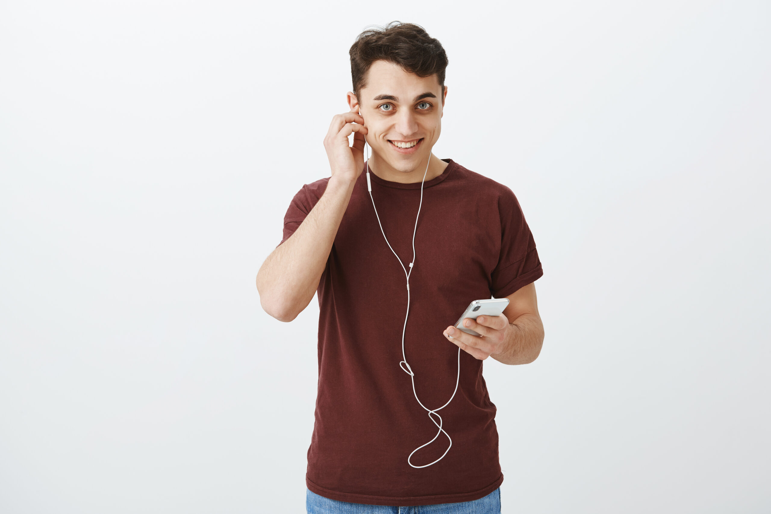 Indoor shot of charming joyful european man in red t-shirt, putting on earphones and holding smartphone, starting listening audio course or music, gazing satisfied and happy over gray background.