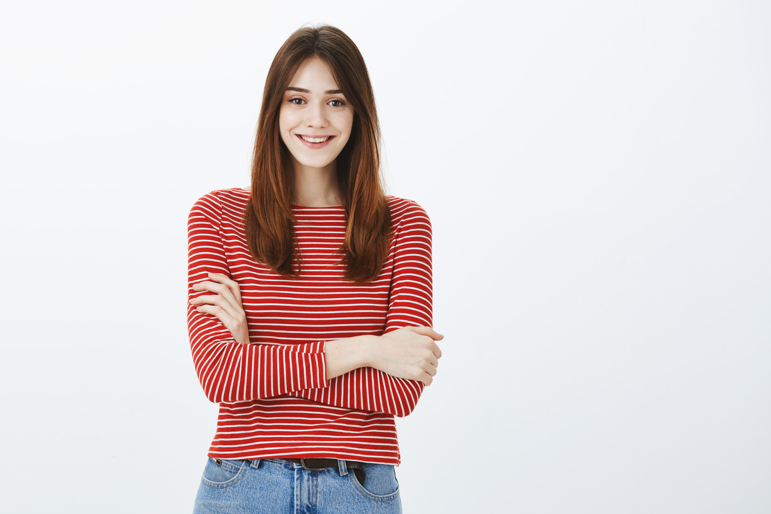 Indoor shot of positive charming european woman with brown hair, holding hands crossed over chest and smiling broadly, feeling uncomfortable but polite over gray background, talking with client.
