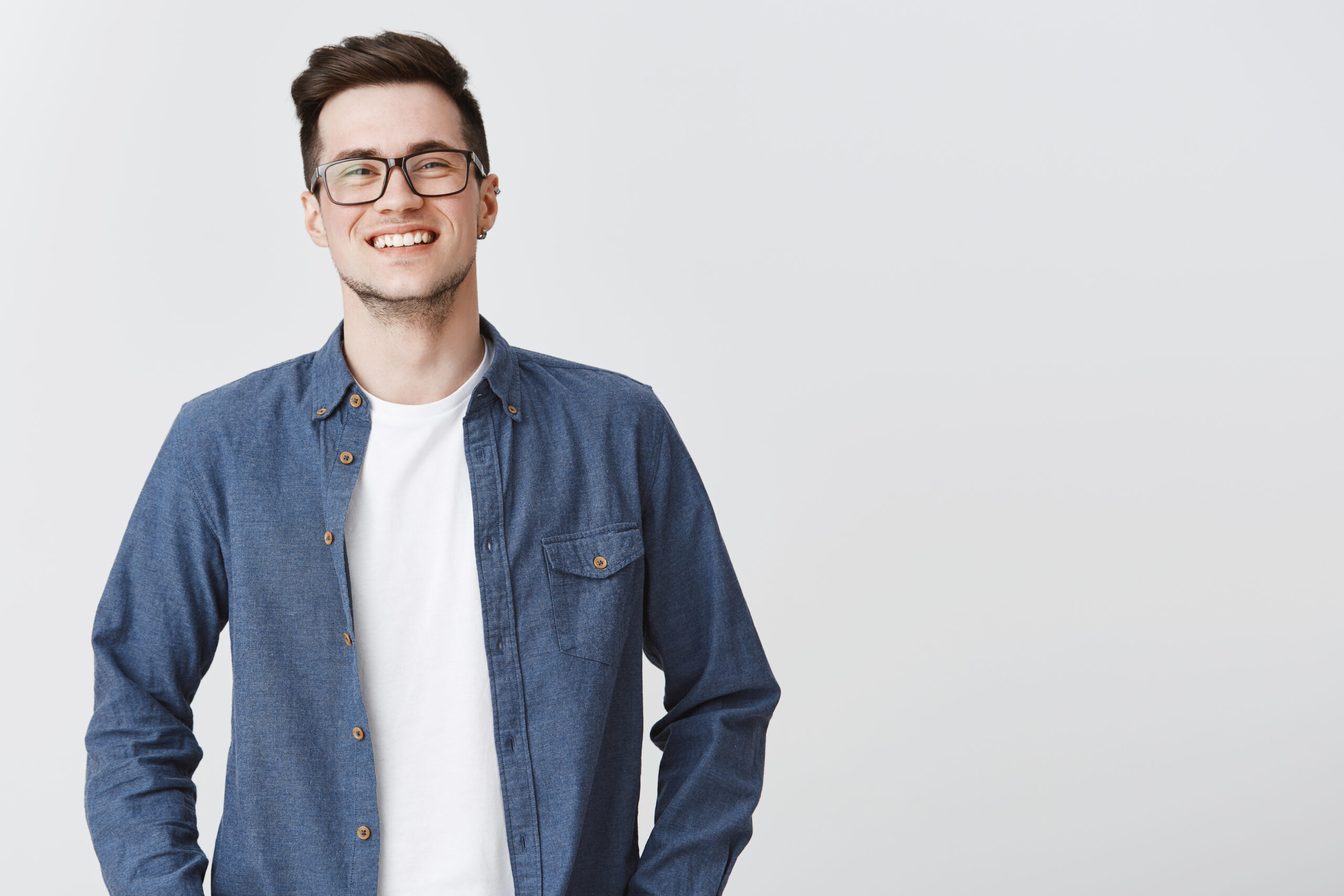 Waist-up shot of happy friendly-looking handsome young smart male coworker in glasses and blue shirt smiling standing over gray background proud and satisfied, grinning at camera from joy. Emotions concept